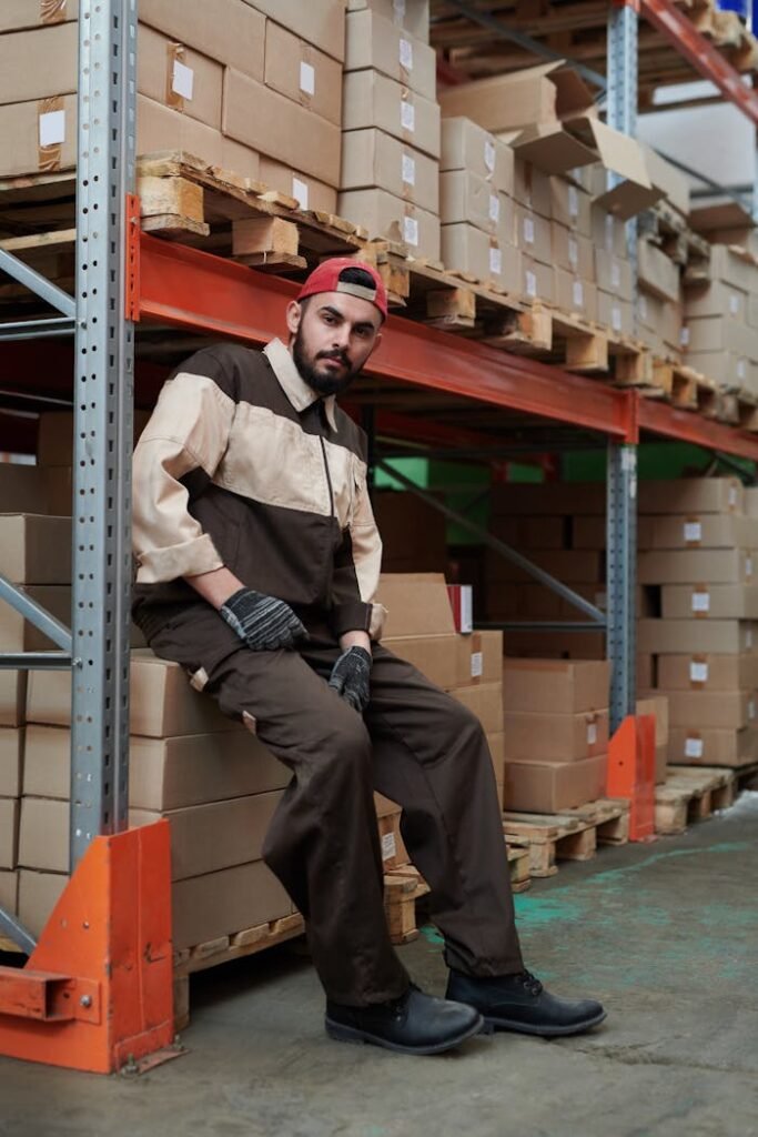 Worker in a warehouse sitting on boxes, wearing a uniform and cap, surrounded by shelves of stock.